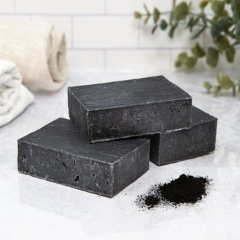 Poofy Organics activated charcoal facial soap bar, natural bath product with black soap and lathered texture in a close-up shot.