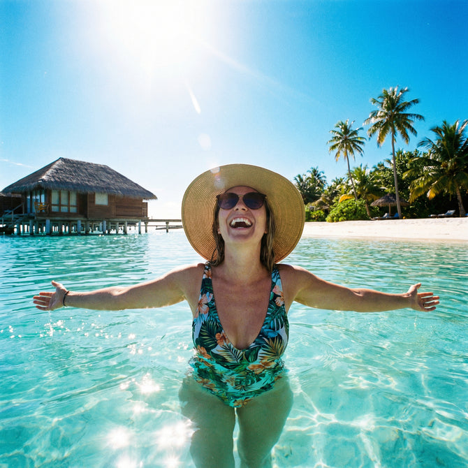Woman in a swimsuit and sun hat standing in clear blue water with palm trees and a hut in the background.